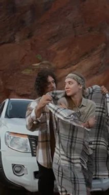 Genç beyaz bir çiftin çölde araba yolculuğu sırasında kamyonun önünde durup kız arkadaşını sıcak bir battaniyeye sarılıp kucaklaşırken orta boy dikey görüntüsü.