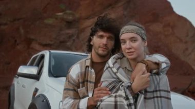 Beyaz kamyonetin yanında durup tatilde çölde dinlenirken kıvırcık saçlı, kız arkadaşına sarılıp battaniyeye sarmalayan beyaz bir adamın orta boy portresi.