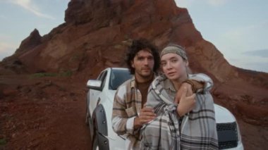 Beyaz kamyonete yaslanan beyaz erkek ve kadının orta boy portresi vahşi doğada duraklarken, sarılırken, romantik alacakaranlık gökyüzüne bakarken.