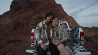 Kamyonet bagajında oturan, kucaklaşan, çay içen, kameraya bakan ve tatilde kırsal alanda yalnız seyahat eden beyaz bir çiftin orta boy portresi.