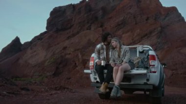 Geniş açıda beyaz erkek ve kadının kamyon bagajında oturup, sarılıp, alacakaranlık gökyüzünde gün batımını seyrederken, balayı ya da tatil sırasında taşlı çölde tek başına seyahat ederken.