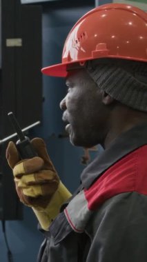 Dikey göğüs hizasında, fabrika işçileri ile telsiz aracılığıyla iletişim kuran, fabrika imalat fabrikasında çalışma saatleri boyunca emirler veren siyah erkek teknisyenlerin kasketli ve tulumlu görüntüleri.
