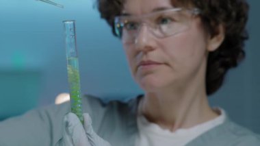 Gözlüklü beyaz kadın bilim adamının yakın çekimi, eldivenler deney tüpündeki yeşil alg iplikçiklerine besin sıvısı damlatırken biyoloji laboratuarında deney yapıyor.