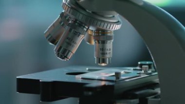 Laboratuvarda mikroskobun yakın plan görüntüsü, cam kaydıraklı eldiven kaldırma masasında çalışan bir araştırmacının eliyle bulanık mavi ve gri arkaplanda objektif bir göz merceği oluşturması.