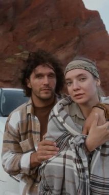 Beyaz araba kaputunda oturan genç beyaz kız ve adamın orta boy dikey portresi vahşi çölde duraklarken, sarılıp kameraya bakarken, alacakaranlıkta