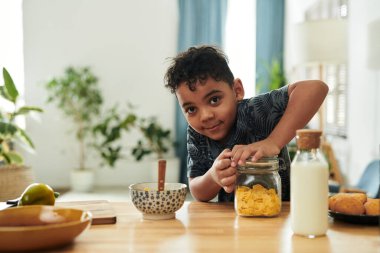 Çizgili tişörtlü şirin çocuk kameraya bakıyor ve kavanozun kapağını mısır gevreğiyle açıyor.