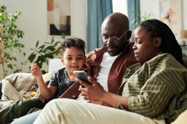 Tatlı küçük çocuk ve babası birlikte vakit geçirirken Afro-Amerikan bir kadının elinde tuttuğu akıllı telefon ekranına bakıyorlar.