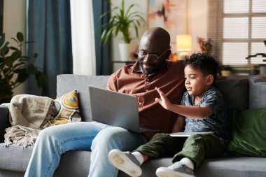 Sevimli küçük bir çocuk dizlerinde bilgisayarla babasının yanında otururken annesine ya da arkadaşına el sallıyor.