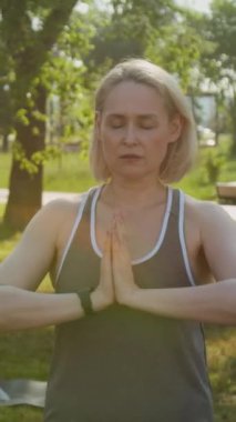 Orta yaşlı, orta yaşlı, dışarıda yoga yapan, çimlerde gözleri kapalı duran ve sıcak yaz gününde namaste işareti yapan beyaz bir kadının dikey fotoğrafı.