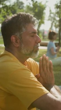 Parlak sarı tişörtlü yaşlı çiftçi adamın, güneşli bir yaz günü açık hava grup çalışması sırasında elleri namaste için birleştirilmiş parktaki çimenlikte gözleri kapalı meditasyon yaparken dikey pozu.