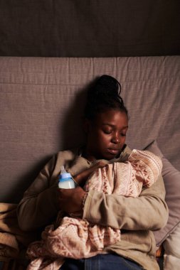 Yeni doğmuş bebeğini sarmalayıp elinde bir şişe sütle yalnız bir anne kameranın önünde koltukta oturup bebek gibi dinleniyor.