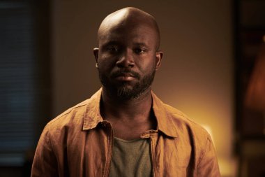 Genç, kel, ciddi, sakallı, tartışmalar sırasında evde dikilirken kameraya bakan Afrikalı Amerikalı bir adam.