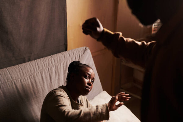 Young miserable African American woman sitting on couch and defending from her aggressive husband holding punch over her