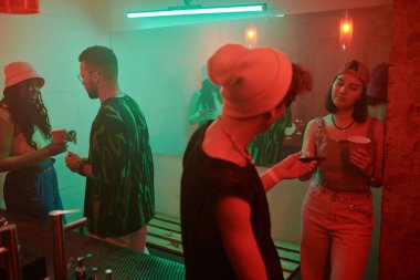 Şık giyinmiş bir adam, gece kulübünde iyi vakit geçirirken kız arkadaşına bir bardak soda ya da başka bir içkiyle cep telefonu uzatıyor.