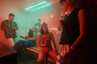 Elinde bira şişesi olan bir adam tuvalette genç ve kültürler arası bir çiftle sohbet ederken kız arkadaşına bir bardak sodayla bakıyor.