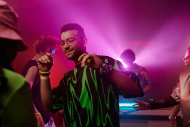Siyah-yeşil tişörtlü, mor neon ışıklı gece kulübünde arkadaşlarına ve DJ 'lere karşı dans eden çok ırklı genç bir adam.