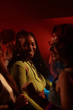 Şık giyinmiş, kültürler arası mutlu insanlar arasında dans eden ve gece kulübünde kırmızı ışıkla eğlenen neşeli genç kız.