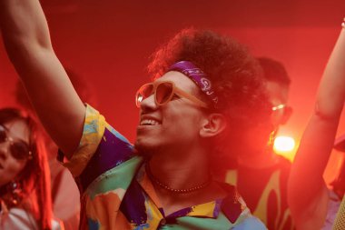 Şık saç bandı ve güneş gözlüğü takan neşeli genç adam gece kulübündeki arkadaşları arasında dans ederken kolunu kaldırıyor.