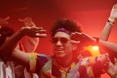 Funk kıyafetli mutlu genç adam gece kulübünde arkadaşlarının arasında dans ederken elini kaldırıyor.