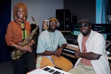 Modern kayıt stüdyosunda çalışırken gülümseyen üç Afrikalı Amerikalı müzisyen yüksek teknoloji müzik aletleri ve enstrümanlarla çevrili.