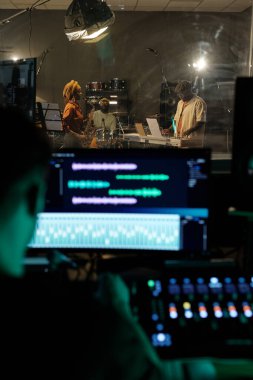 Musicians performing in recording studio with sound engineer managing control panels. Visible instruments and studio equipment in background, showcasing music production environment clipart