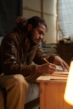 Samimi sanat stüdyosunun ahşap masasında otururken çizim yapmaya konsantre olan adam çalışma alanını aydınlatıyor.