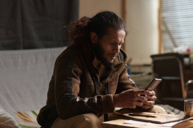 Uzun saçlı ve sakallı bir adam evde rahat bir şekilde oturup kitap okuyor. Basit bir dekor ve doğal ışıkla.