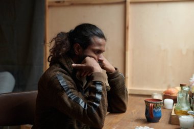 Düşünceli genç adam ahşap masada kırsal mutfak ortamında oturuyor, düşünceli bir ifadeyle çenesini dinlendiriyor, çeşitli baharatlar ve kavanozlarla sıcacık bir şekilde aydınlatıyor.