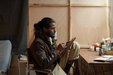 Uzun saçlı ve sakallı bir adam, elinde kalem ve defter tutarak oturmuş, derin düşüncelere dalmış, yazma ya da çizim yaparken, doğal ışıkla iç mekan sahnesi