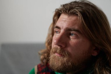 Uzun saçlı ve sakallı bir adam içeride oturmuş, düşünceli bir şekilde uzaklara bakıyor. Yumuşak doğal ışık yüzünü aydınlatıyor, derin düşünme havasını yansıtıyor.