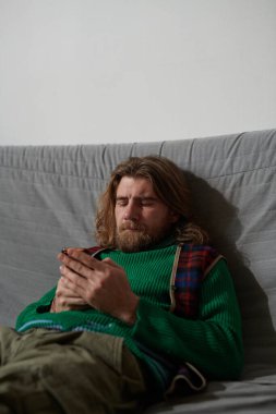 Yeşil kazak ve ekose eşarp giyen sakallı adam kanepede oturuyor ve telefonda yazıyor, kameraya bakmadan odaklanmış yüz ifadesi sergiliyor.