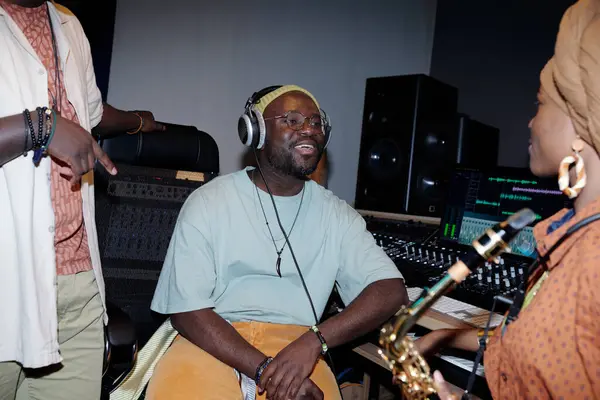 stock image Man smiling while wearing headphones in a recording studio filled with music equipment. Other individuals interacting with him, creating a lively atmosphere