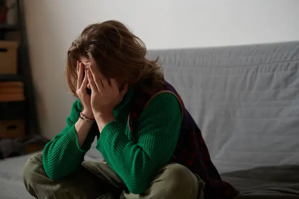 stock image Young man seated on gray couch hiding face with hands in a candid moment revealing long hair and casual attire creating a sense of introspection