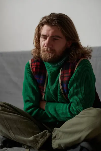 stock image Man with shoulder-length hair and beard sitting cross-legged on a couch with arms crossed wearing green sweater and plaid vest