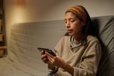 Hispanic woman sitting on sofa using smartphone while surrounded by dimly lit cozy room, wearing casual clothes and focused on screen clipart