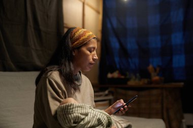 Konsantre bir kadın, arka planda mavi ekose perde ile loş ışıklı odada akıllı telefon kullanırken koltukta oturuyor, çizgili gömlek ve saç bandı takıyor, elinde fincanı başka bir elde tutuyor.