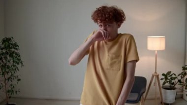 Hardal tişörtlü genç beyaz adamın orta boy portresi tiksinti duyuyor, modern minimalist odada suratını buruşturuyor, kameraya bakıyor.