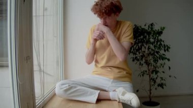 Tişörtlü ve pantolonlu beyaz bir adam ahşap pencere eşiğinde oturmuş, iğrenç manzaraya bakıyor, midesi bulanıyor.