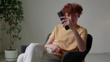 Z jenerasyonunun beyaz tenli, günlük kıyafetlerini giymiş uzaktan kumandayla televizyon kanallarını değiştirip evde film seçen orta boy fotoğrafı.