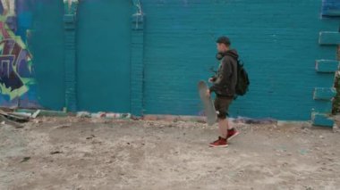 Beyaz erkek grafiti sanatçısı arka sokaktaki mavi tuğladan duvara doğru yürürken, kaykayı indirirken, resim yerini incelerken, bir yandan da başyapıt yaratmaya hazırlanırken.