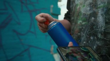 Ekranda yeni etiket tasarımı olan akıllı telefonu tutan ve boya şişesindeki sprey başlığı değiştiren isimsiz erkek graffiti sanatçılarının elleri, duvarda resim yapmaya hazırlanırken,