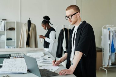Boyunda mezurası olan genç ciddi bir terzi dizüstü bilgisayara bakıyor ve notlar alıyor ya da yeni koleksiyon için çizimler yapıyor.