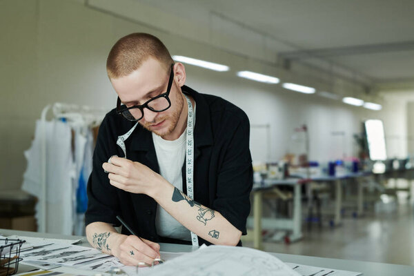 Young serious couturier or tailor with measuring tape on neck bending over workplace in atelier and sketching or making notes in notepad