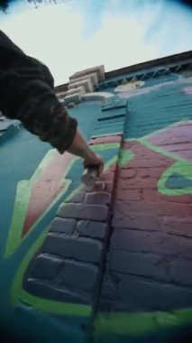 Gaz maskeli, kazaklı, yeşil sprey boyayla kırmızı-mor grafitilerin etrafını çizen, kamu malına zarar veren genç bir adamın el kamerası görüntüsü.