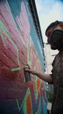 Arka sokaktaki tuğla duvarı dekore ederken solunum maskesi takan, kırmızı-mor arka planda yeşil sprey boya ile etiketli erkek grafiticinin düşük açılı dikey görüntüsü.
