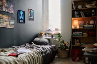 Duvarda bilim kurgu posterleri, yastıklı temiz bir yatak odası ve çeşitli kitap ve dekorasyonlarla dolu ahşap bir kitaplık bulunan modern yatak odası. Lambadan gelen sıcak ışık davetkar atmosfer yaratıyor