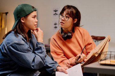 İki kadın sanat stüdyosunda sohbet ediyor, biri elinde kağıt ve kalemle, ikisi de günlük kıyafetler içinde. Arka planda çeşitli sanat malzemeleri ve çizimler yer alıyor