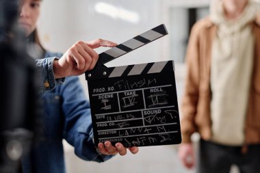 Film setindeki bir film sahnesine hazırlanırken, ekip üyesi elinde bir alkış tutuyor. Bulanık arka plan, film yapım sahnesine derinlik katan ayakta duran başka bir ekip üyesini içeriyor.