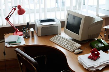 Bilgisayarı, yazıcıyı, kırmızı lambayı, yeşil klasörü, kalem bardaklarını, not defterlerini ve zımbayı gösteren düzgün düzenlenmiş ofis masası. Doğal ışık sağlayan büyük pencere çalışma alanını aydınlatır