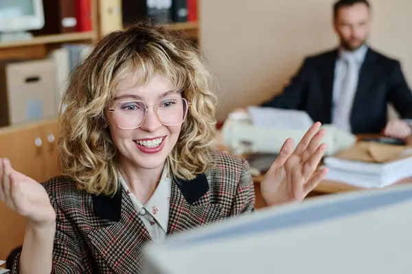 stock image Smiling woman gesturing enthusiastically while sitting at desk in office environment with colleague working in background, capturing excitement and positive vibes